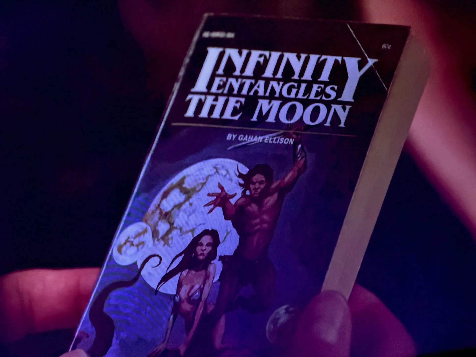 A worn copy of Infinity Entangles the Moon held in purple and blue light. The photo was taken of my TV during an episode of The Rookie.